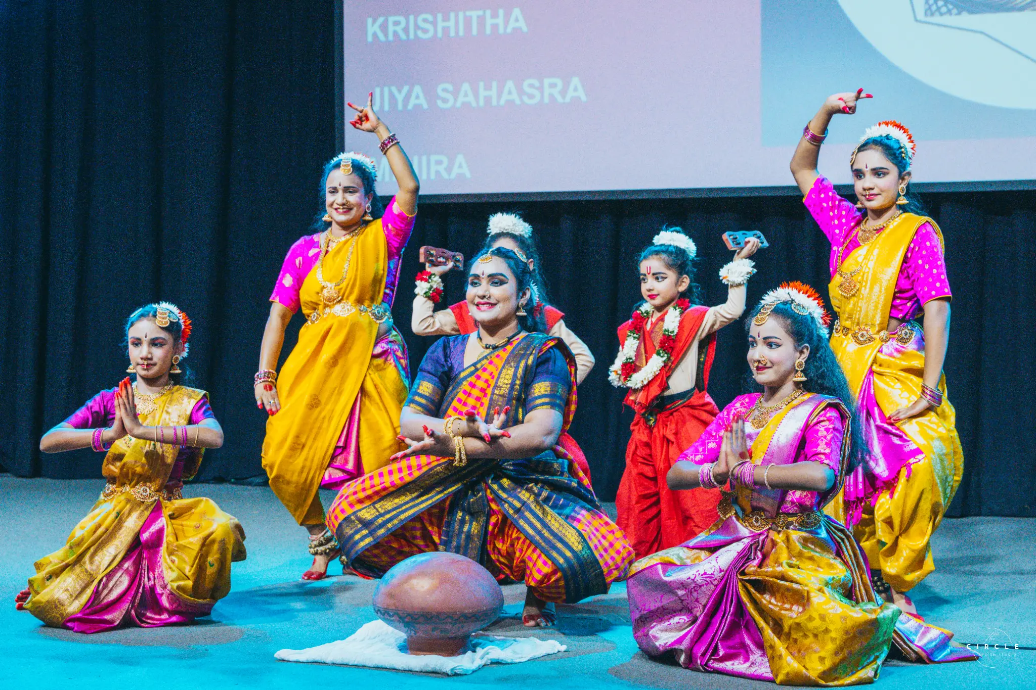 Six performers in colourful costumes on a blue-lit stage at a Melbourne event